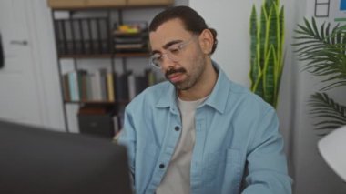 Ofiste gözlerini ovuşturan genç adam bilgisayarın önünde yorgun, gözlüklü ve mavi gömlekli.