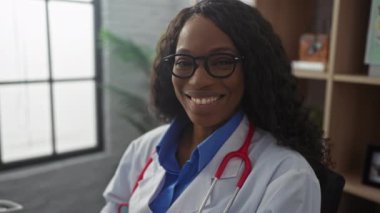 Gözlüklü, kıvırcık saçlı, kendine güvenen Afro-Amerikan kadın doktor modern bir klinikte beyaz önlük ve steteskop giyerek otururken gülümsüyor..