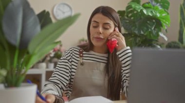 Çizgili tişörtlü, odaklanmış bir İspanyol kadın bitkilerle çevrili bir evde çalışırken telefon görüşmesi yapıyor..
