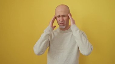 Sessizlik içinde acı çeken, çaresiz, üzgün, sakallı, kazak giyen, stres kaynaklı migren ağrısıyla başa çıkan genç bir İspanyol. Yapayalnız, el üstünde, izole edilmiş sarı arka planda.