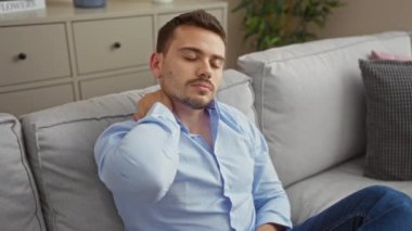 Mavi gömlekli yakışıklı İspanyol bir adam evdeki kanepede oturuyor, rahat olmayan boynunu tutuyor, modern dekorlu rahat bir oturma odasında..