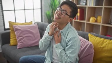 Genç Asyalı bir adam modern oturma odasındaki bir kanepede otururken boynunu kaşıyarak rahatsızlık ya da kaşıntı yaratıyor..