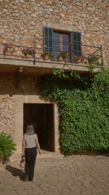 Genç bir kadın güneşli bir günde Mallorca, İspanya 'da geleneksel taş ev girişini keşfediyor. Akdeniz mimarisinin ve yaşam tarzının özünü canlı yeşillik ve kırsal cazibeyle yakalıyor.