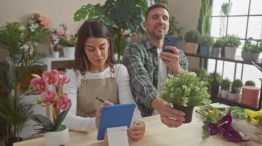 Bir kadın ve erkek, bitkilerle çevrili bir çiçekçide iş birliği yapıyor, notlar alıyor ve akıllı telefon kullanıyor, takım çalışması ve küçük işletmeler sergiliyor.