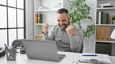 Sakallı ve gri saçlı olgun İspanyol adam modern bir ofiste başarıyı kutluyor..