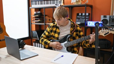 Masasında dizüstü bilgisayarı ve kulaklığı olan bir müzik stüdyosunda gitar akort eden sarışın bir adam..