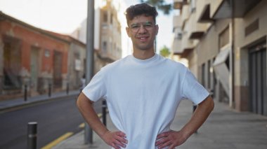 Genç, yakışıklı İspanyol bir adam, güneşli bir günde şehrin arka planında, gözlüklü ve beyaz tişörtlü, kendine güvenen bir şekilde dikiliyor..