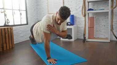 Genç, yakışıklı İspanyol bir adam mavi bir paspasın üzerinde spor yapıyor. Kapalı bir spor salonunda modern, iyi donanımlı bir spor salonu sergiliyor..