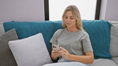 Çekici genç bir kadın turkuaz bir kanepede dinleniyor, modern bir oturma odasında akıllı telefonunu karıştırıyor..