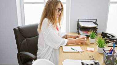 Masasında çalışan genç bir kadın. Parlak bir ofis odasında bir fincan ile..