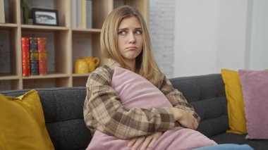 Rahat bir oturma odasında yastıkla kanepede oturan üzgün genç bir kadın, başka tarafa bakıyor..