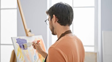 Sakallı genç bir adam parlak bir stüdyo içinde tuvale resim yapıyor, yaratıcılık ve odaklanma sergiliyor..