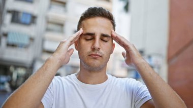 A stressed young hispanic man outdoors in a city, portraying a headache with his fingers on temples. clipart