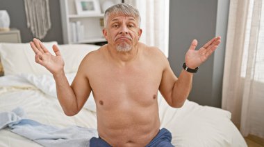 Gömleksiz, kafası karışık, orta yaşlı bir adam. Kapalı bir yatakta oturuyor ve omuz silkiyor..