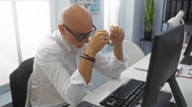 Gri saçlı, sakallı ve gözlüklü bir adam ofis masasında çalışıyor. Parlak ışıklandırılmış, kapalı bir ortamda. Evrak işlerine odaklanıyor.