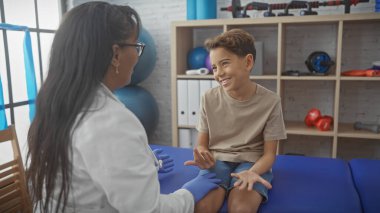 Bir kadın terapist, gülümseyen bir çocuğun bacağını iyi donanımlı bir fizyoterapi odasında muayene eder..