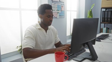 Modern ofis odasında bilgisayar başında çalışan yakışıklı Afro-Amerikalı adam. Parlak pencereleri ve düzenli çalışma alanı var.