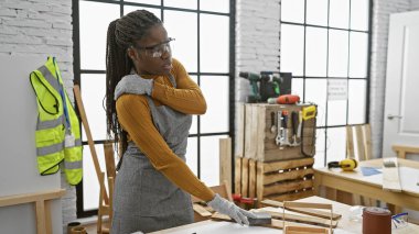 Örgü ören siyah bir kadın marangozluk atölyesinde sırt ağrısı çekiyor, güvenlik gözlüğü takıyor ve günlük giysiler giyiyor..