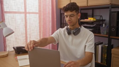 Ofis ortamında dizüstü bilgisayarda çalışan, gözlük ve kulaklık takan, raflı ve arka planda organize belgeler olan genç İspanyol bir adam..