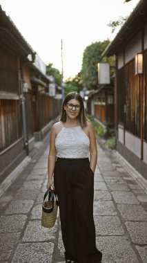 Kendine güvenen İspanyol kadın, güzel ve neşeli, gözlükleriyle neşeli poz veren, Kyoto 'nun geleneksel sokaklarında ayakta duran ve gülümseyen, doğal ve kaygısız Latin ifadesi yayan..