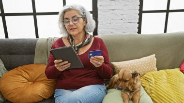 Gözlüklü, elinde kredi kartı ve tablet olan olgun bir kadın. Yanında köpek var. Kapalı koltukta dinleniyor..
