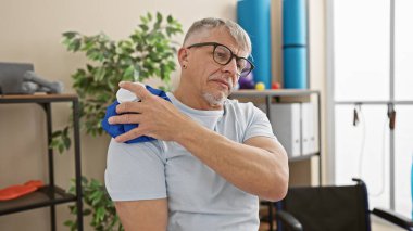 Mature grey-haired man applies ice pack to shoulder in therapy room, showing pain management. clipart