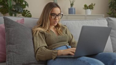 Genç, çekici, gözlüklü sarışın bir kadın modern bir evde rahat bir koltukta otururken dizüstü bilgisayar kullanıyor..