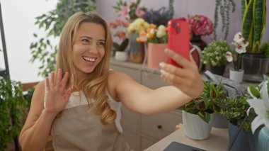 Gülümseyen sarışın kadın çiçekçi dükkanında telefonla selfie çekiyor..