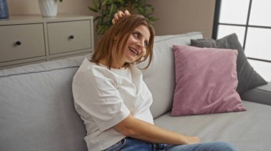 Genç, çekici, beyaz bir kadın evdeki rahat bir oturma odasında rahat bir kanepede dinlenirken gülümsüyor..