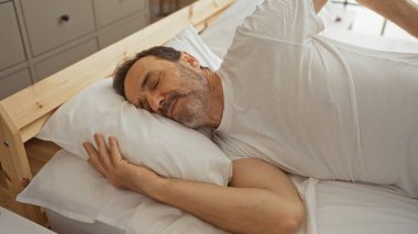 Yakışıklı İspanyol adam rahat bir yatak odasında huzurlu bir şekilde uyuyor. Parlak bir iç mekanı var. Rahat bir evde huzurlu bir sabah sergiliyor..