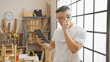 Genç İspanyol bir adam marangozluk atölyesinde akıllı telefon ve tabletle birden fazla iş yapıyor..