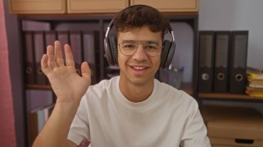 Modern ofis alanındaki bir video konferansında gülümseyen ve el sallayan gözlüklü İspanyol genç adam..