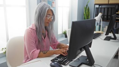 Ofiste klavyeye yazı yazan bir kadın, olgun, gri saçlı, kapalı alanda çalışan ve profesyonel bir odada bilgisayara odaklanan bir kadın.