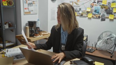 Odaklanmış sarışın bir kadın dedektif, dava dosyaları ve ilan panosuyla çevrili bürosundaki kanıtları analiz eder..