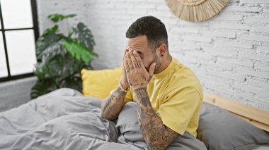 Dövmeleri olan sıkıntılı bir İspanyol adam yüzünü ellerle örterek beyaz tuğla kaplı bir yatak odasında oturuyor..