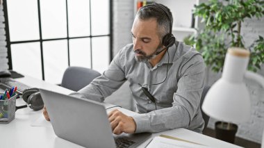 Parlak modern ofis ortamında dizüstü bilgisayarda çalışan kulaklıklı İspanyol bir kıdemli adam.