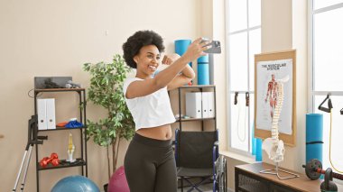 Arka planda spor aletleri olan modern bir spor salonunda selfie çekerken gülümseyen Afrikalı Amerikalı kadın.