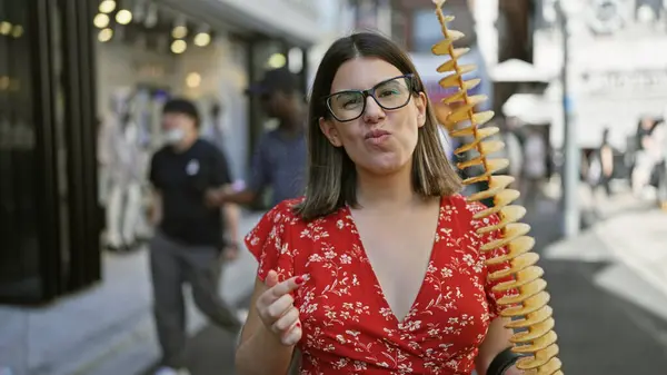 stock image Beautiful hispanic woman joyfully eating crunchy, delicious chips on a stick at takeshita street, tokyo - traveling junk food lover, sporting glasses and a cheerful smile in japan's urban city