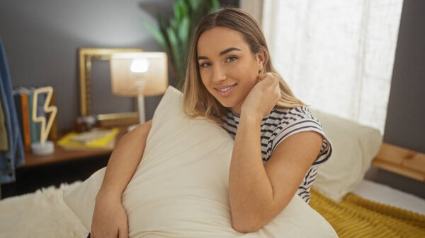 Красивая молодая женщина с светлыми волосами расслабляется в уютной спальне, сидя на кровати, обнимая подушку в стильной обстановке интерьера.