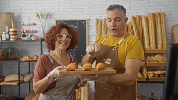 stock image Smiling man and woman bakers working together in a bakery holding a tray of fresh croissants in an indoor shop setting with breads and pastries displayed in the background