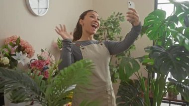 Neşeli bir İspanyol kadın yeşil bitkiler ve renkli çiçeklerle çevrili bir çiçekçide selfie çekiyor..