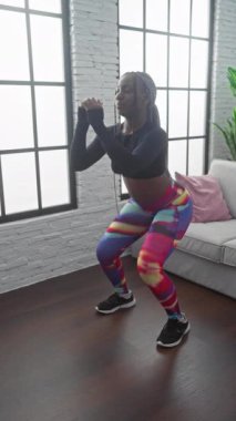 Afrika kökenli Amerikalı kadın içeride egzersiz yapıyor, renkli tayt giyiyor ve siyah bluz giyiyor, modern bir oturma odasında çömeliyor.