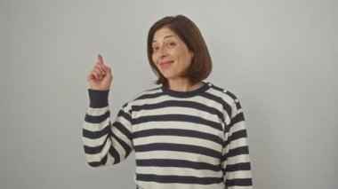 Neşeli, olgun İspanyol kadın, mutlulukla kenarı işaret ediyor! Çizgili bir süveter giyiyor, iyimser gülüşü beyaz, izole bir arkaplanda yayılıyor..