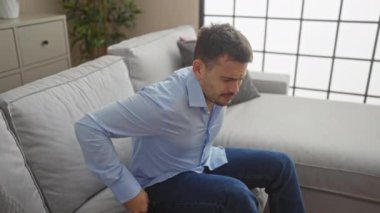 Modern bir oturma odasında sırtı ağrıyan, sakallı, genç bir İspanyol..
