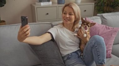Sarışın genç bir kadın oturma odasındaki gri bir kanepede oturmuş kucağında küçük bir chihuahua köpeğiyle selfie çekiyor..
