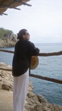 Güneş gözlüğü takan ve elinde hasır şapka tutan gülümseyen kadın Mallorca, İspanya 'daki Cala Deia' dan okyanus manzarasına hayran kalarak kayalık sahil şeridine karşı sakin bir açık hava anı yaşıyor.