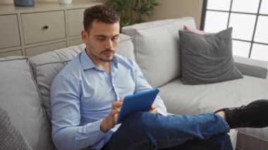 Yakışıklı bir İspanyol, modern bir oturma odasındaki koltukta uzanırken, rahat bir ifade ve günlük giysiler giyer ve tablet kullanır..
