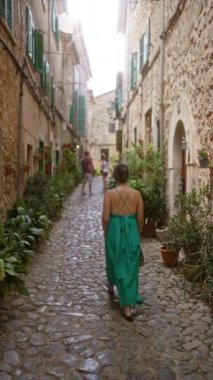 Valldemossa, Mallorca, İspanya 'nın dar bir taş caddesinde yürüyen kadın, güneşli bir günde yeşil bir elbise giyiyor ve etrafında tarihi binalar ve yeşillikler var..