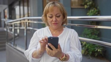 Şehir dışında yaşayan orta yaşlı sarışın bir kadın şehir caddesinde dikilirken akıllı telefonuyla meşgul oluyor. Modern bir bağlantı ve iletişim anını yansıtıyor..