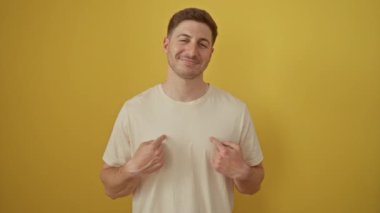 Genç İspanyol adam ayakta ve mutlu, gülümseyen yüz ve parmak ile izole edilmiş sarı arka plan üzerinde duruyor - heyecanlı ve iyimser ifade